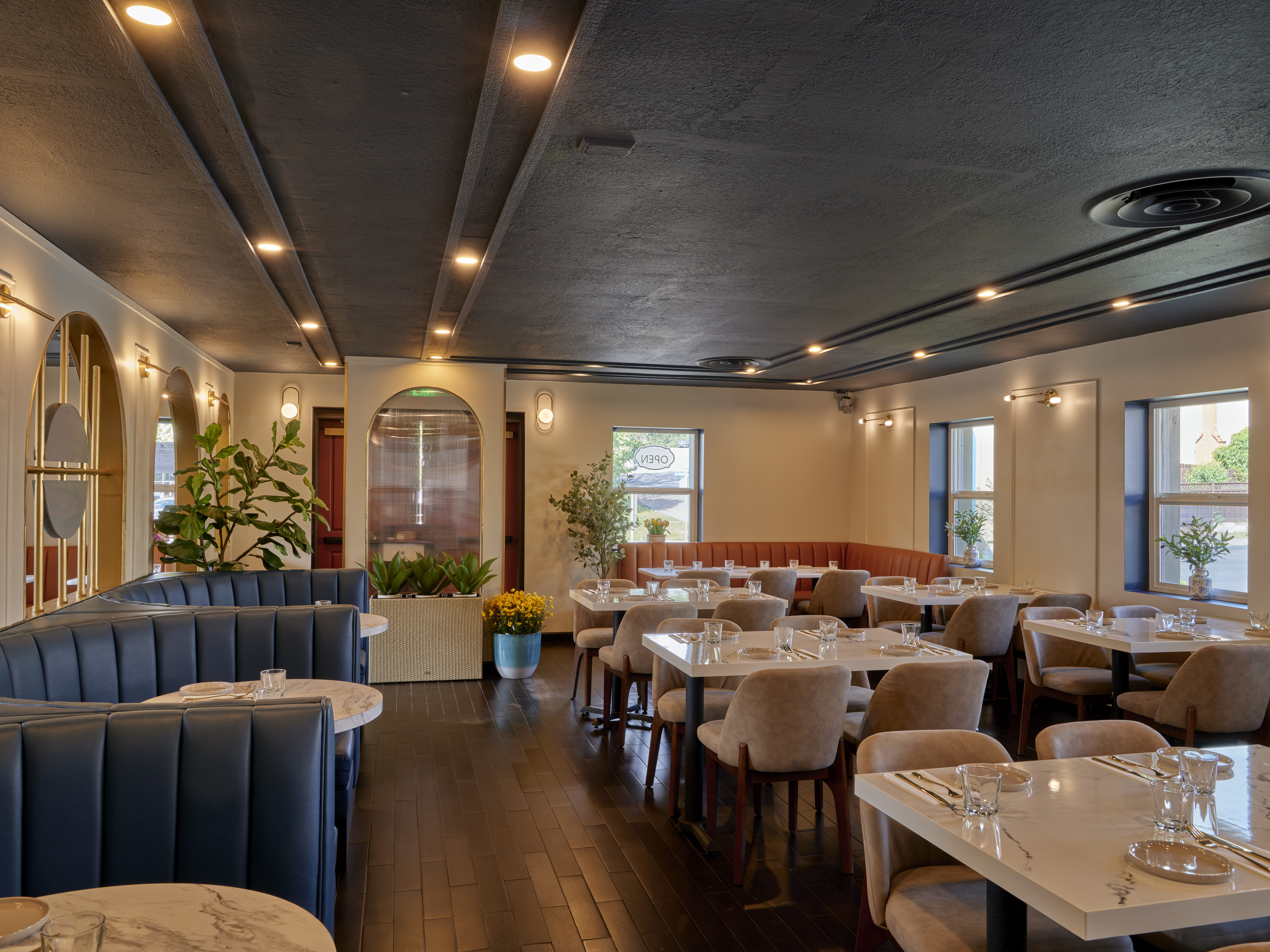 Main dining room at Prettiplatters San Anselmo — navy leather booths, dark ceiling with recessed lighting, white marble tables, and lush greenery